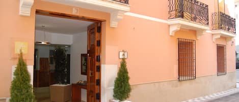 Entrada de hotel con puerta de madera, dos árboles de Navidad en macetas y un balcón con barandillas de metal decorativas.
