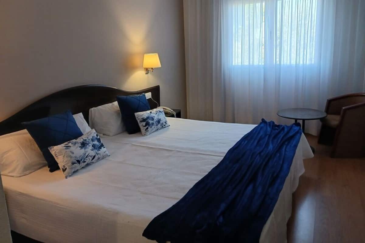 Habitación de hotel con una cama grande, dos almohadas, una lámpara de noche, una silla y una mesita.