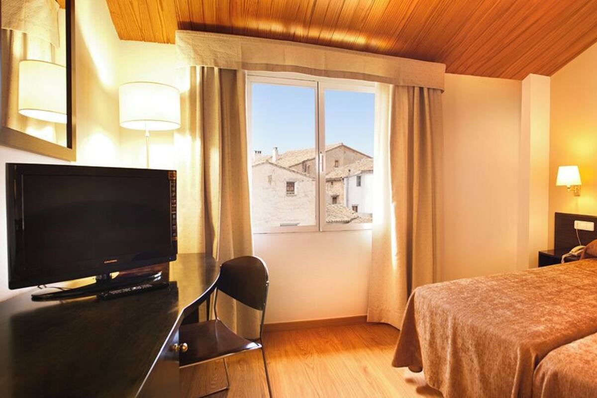 Una habitación de hotel con una cama, un escritorio con televisor, una silla y un ventanal con cortinas.