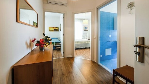 A hallway with wooden flooring, a mirror, a vase with flowers, and a door leading to a room with a bed and a blue wall. - CAROLINA 7 - Lido di Jesolo, Italy. Entire accommodation (Lido di Jesolo)