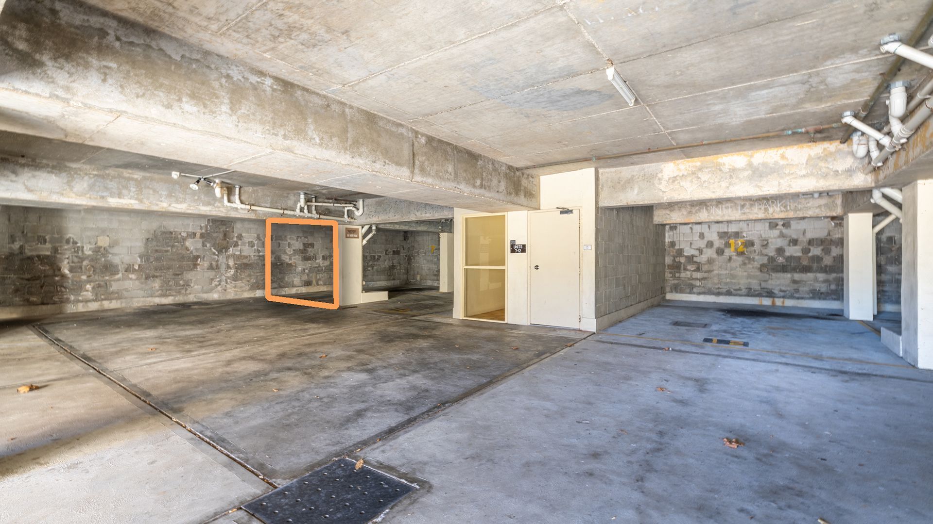 An unfinished parking garage with concrete floors and walls, exposed pipes, and a door labeled "UNITS 7-2".