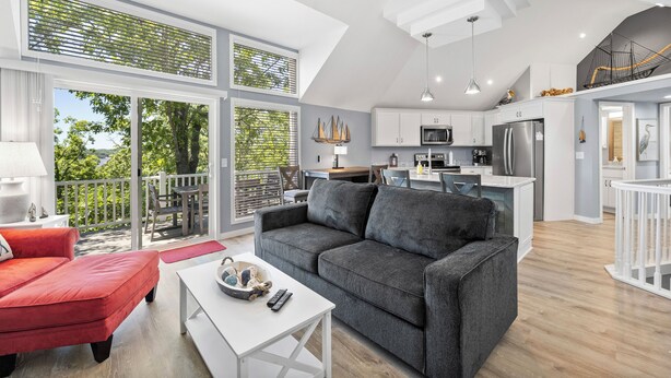 Een ruime woonkamer met een grijze bank, een rode fauteuil en een witte salontafel. Een balkon met uitzicht op groen is zichtbaar.