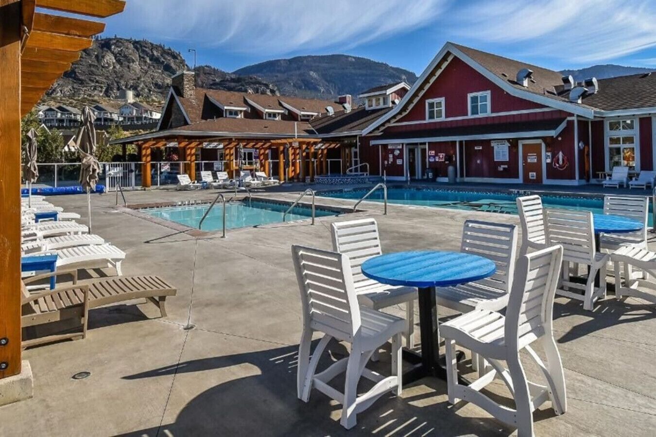 Un espace aménagé au bord de la piscine avec des chaises longues et une table, un bâtiment à la façade rouge et un paysage montagneux en arrière-plan.
