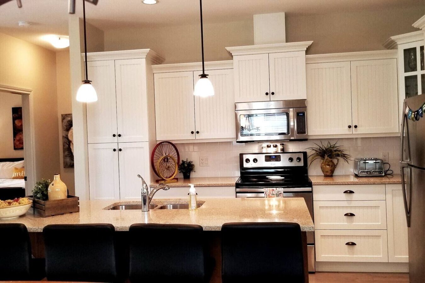A modern kitchen with white cabinets, a stainless steel oven, a microwave, and a refrigerator. There are pendant lights above the island and a large window in the background.