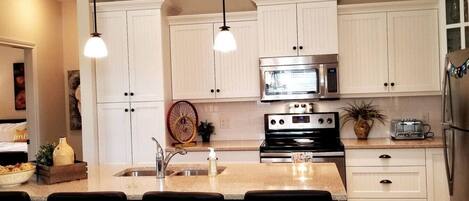A modern kitchen with white cabinets, a stainless steel oven, a microwave, and a refrigerator. There are pendant lights above the island and a large window in the background.