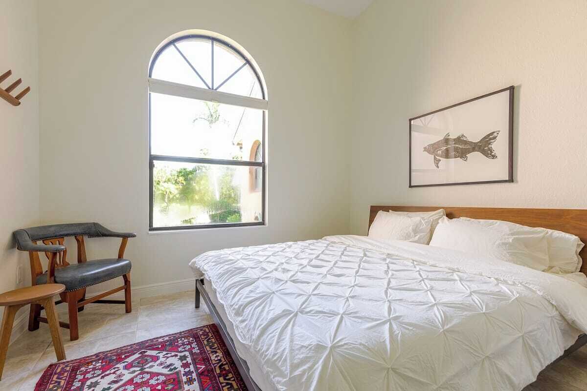 A bedroom with a large window, a bed with white bedding, a wooden bench, and a framed fish picture on the wall.