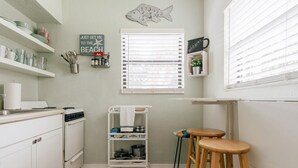 Una cocina con armarios blancos, una estufa, un lavamanos y un taburete de madera. Hay una repisa con vajilla y un cartel que dice "Just get me to the beach".