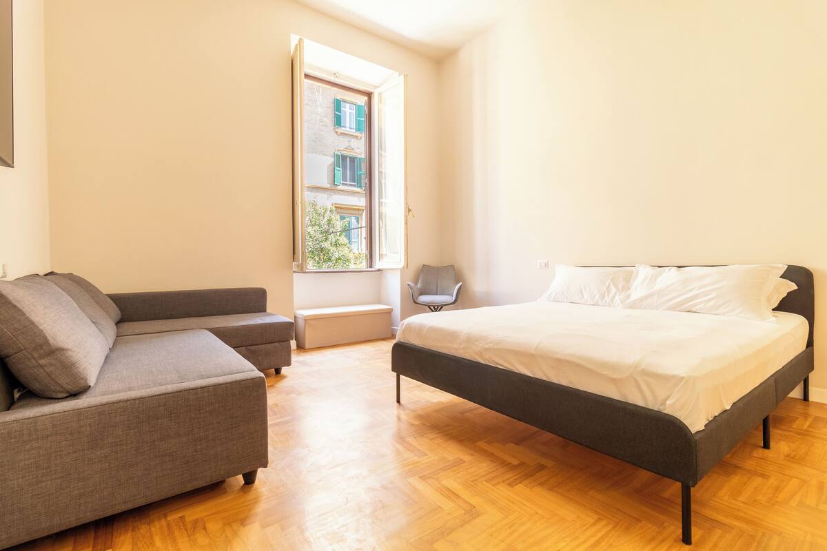 Un dormitorio con cama, sofá, banco y ventana con vistas a edificios.