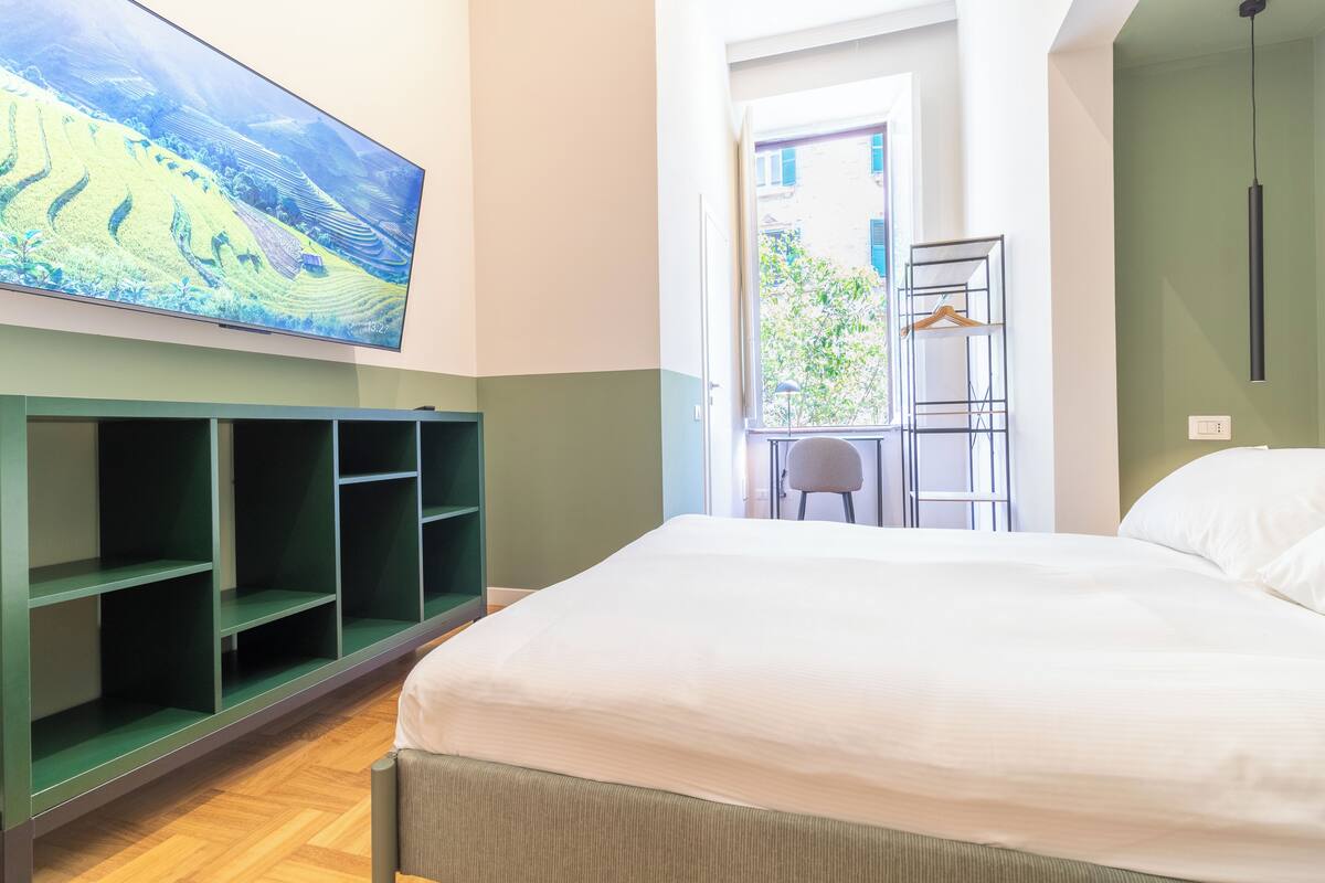 Habitación de hotel con una cama grande, una estantería verde, un televisor en la pared y vistas al exterior.
