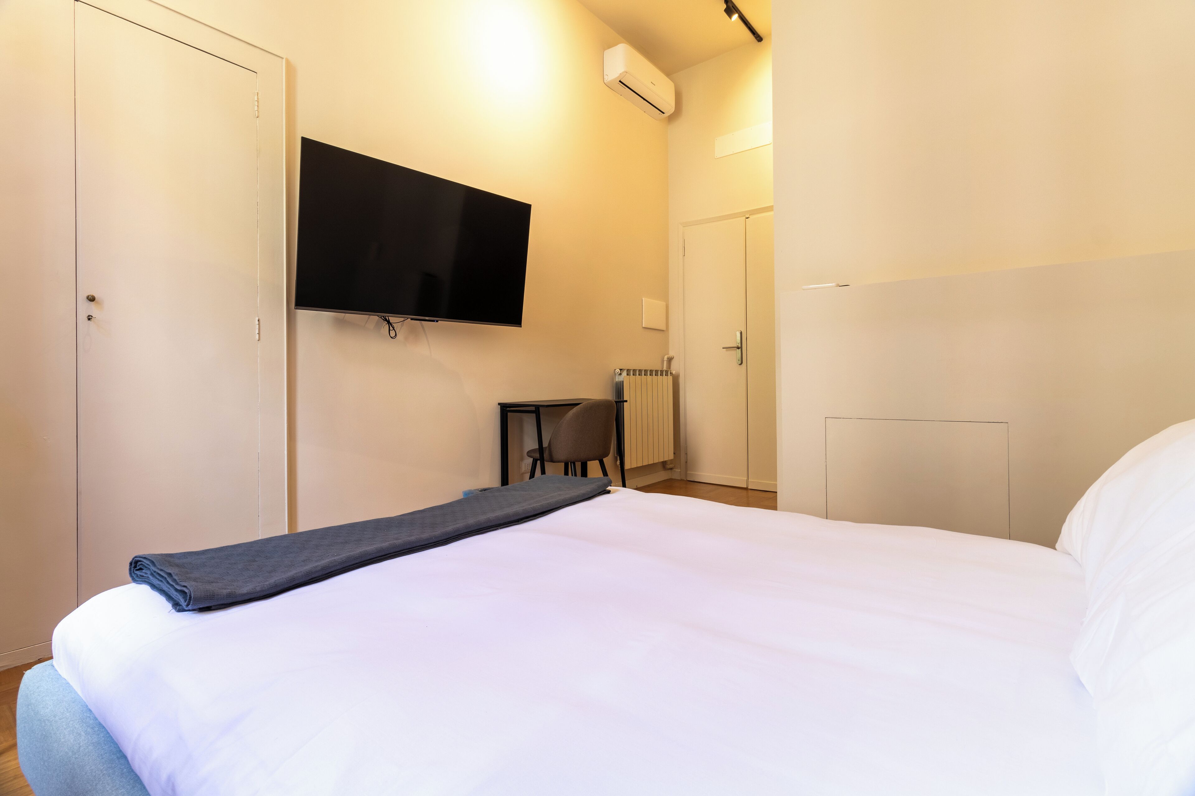 A hotel room with a bed, a TV mounted on the wall, a desk with a chair, and a radiator.