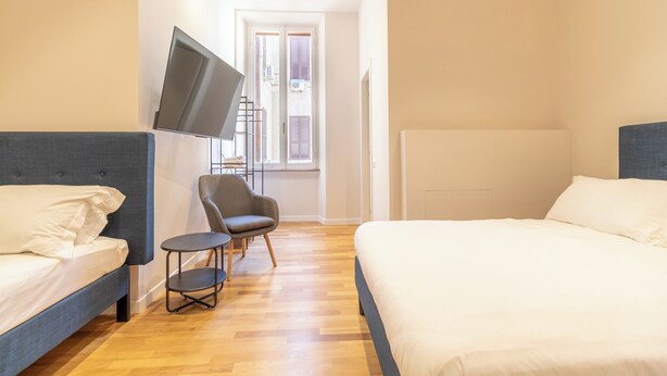 A bedroom with two beds, a TV mounted on the wall, a chair, and a window with shutters. - Cunabu Emilio (Rome)