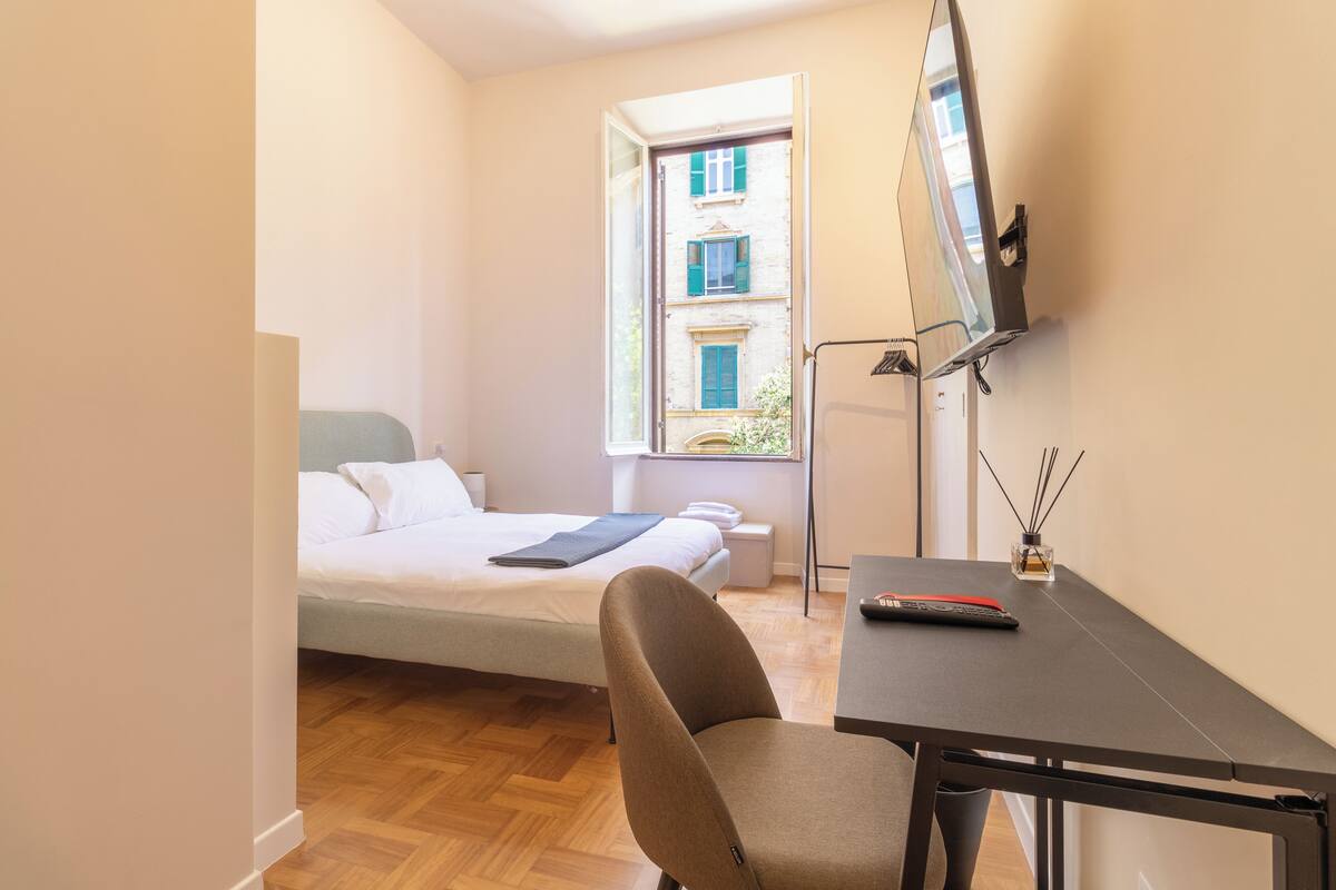 Un dormitorio con cama, escritorio, silla, televisor y ventana con vista a edificios.