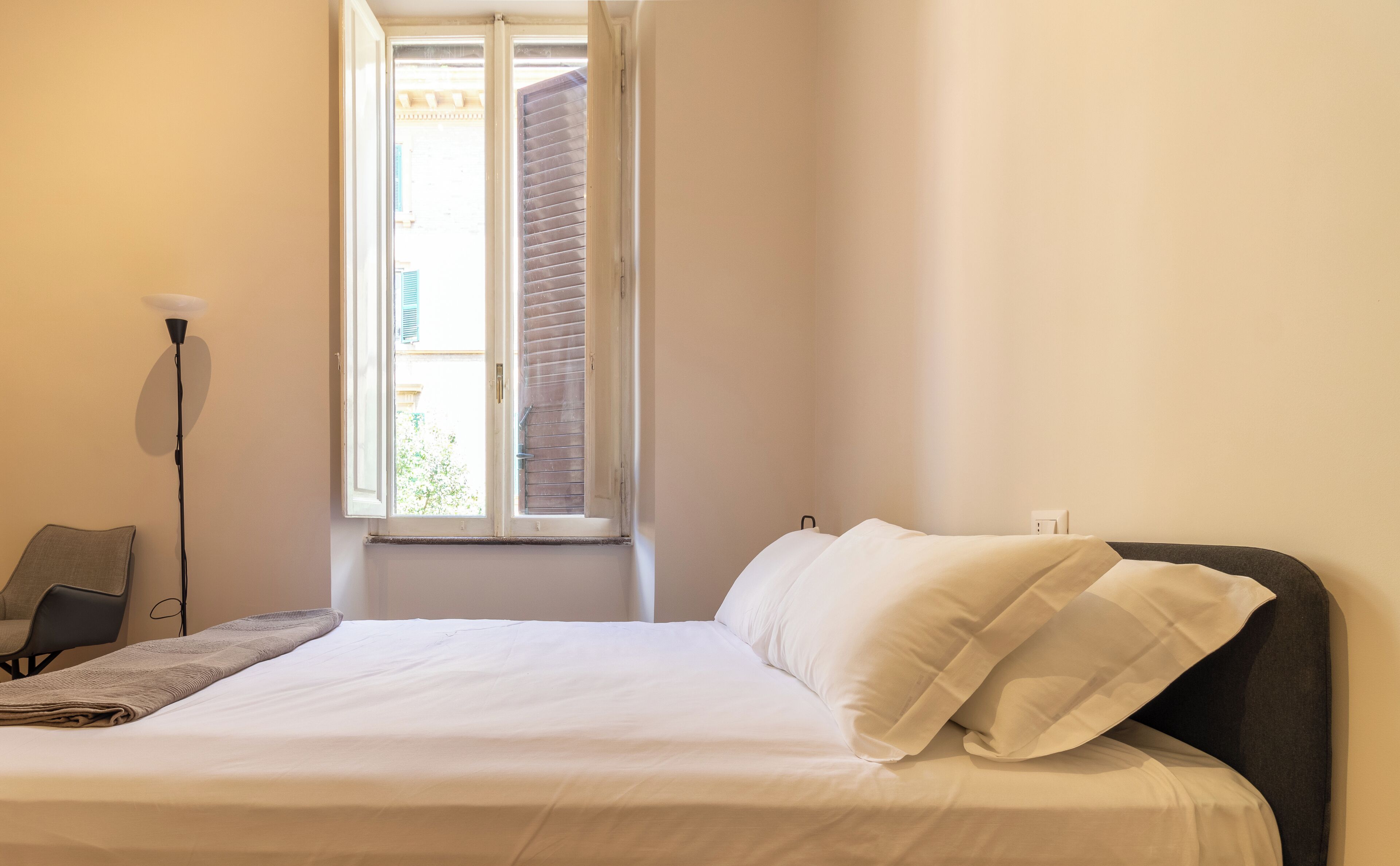 A neatly made bed with white linens in a room with a window, a floor lamp, and a chair.