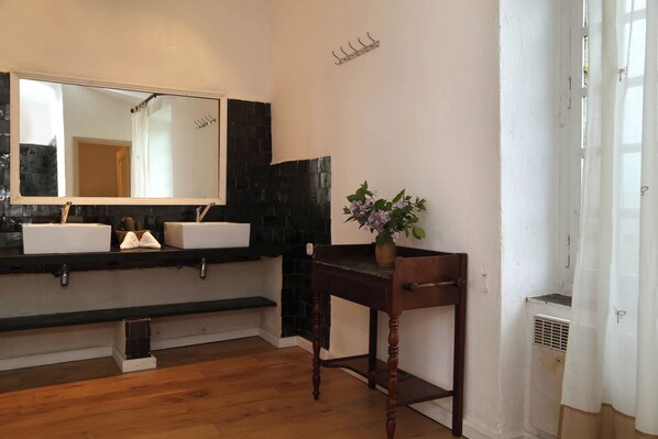 Une salle de bain avec un grand miroir, deux lavabos, une table en bois ornée d’un vase et un radiateur.