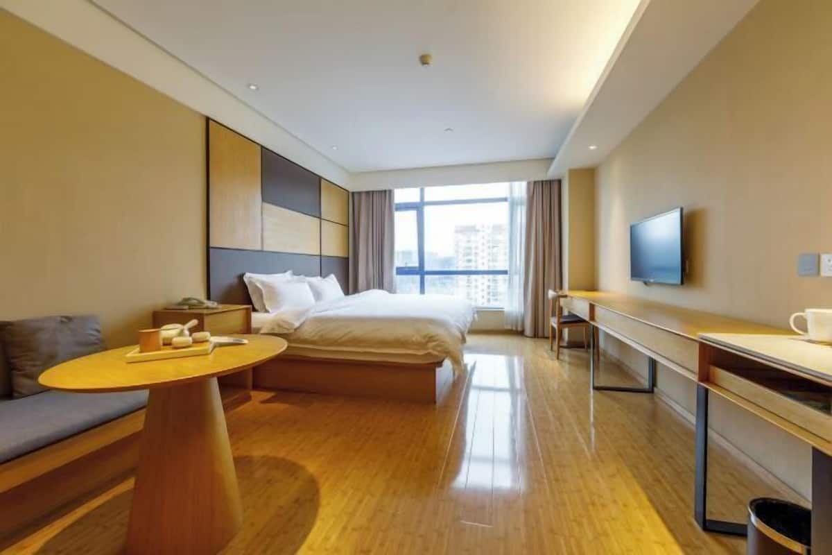 A modern hotel room with a bed, a sofa, a small table, a TV, and a large window offering a city view.