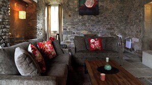 Una habitación rústica con paredes de piedra, dos sofás grises con cojines estampados, una mesa de centro de madera y una obra de arte de granada en la pared.