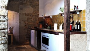 Una cocina rústica con paredes de piedra, una viga de madera, un horno empotrado y un estante con botellas.