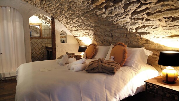 Un dormitorio de paredes de piedra con una cama grande, ropa de cama blanca y una mesita de noche con una lámpara.