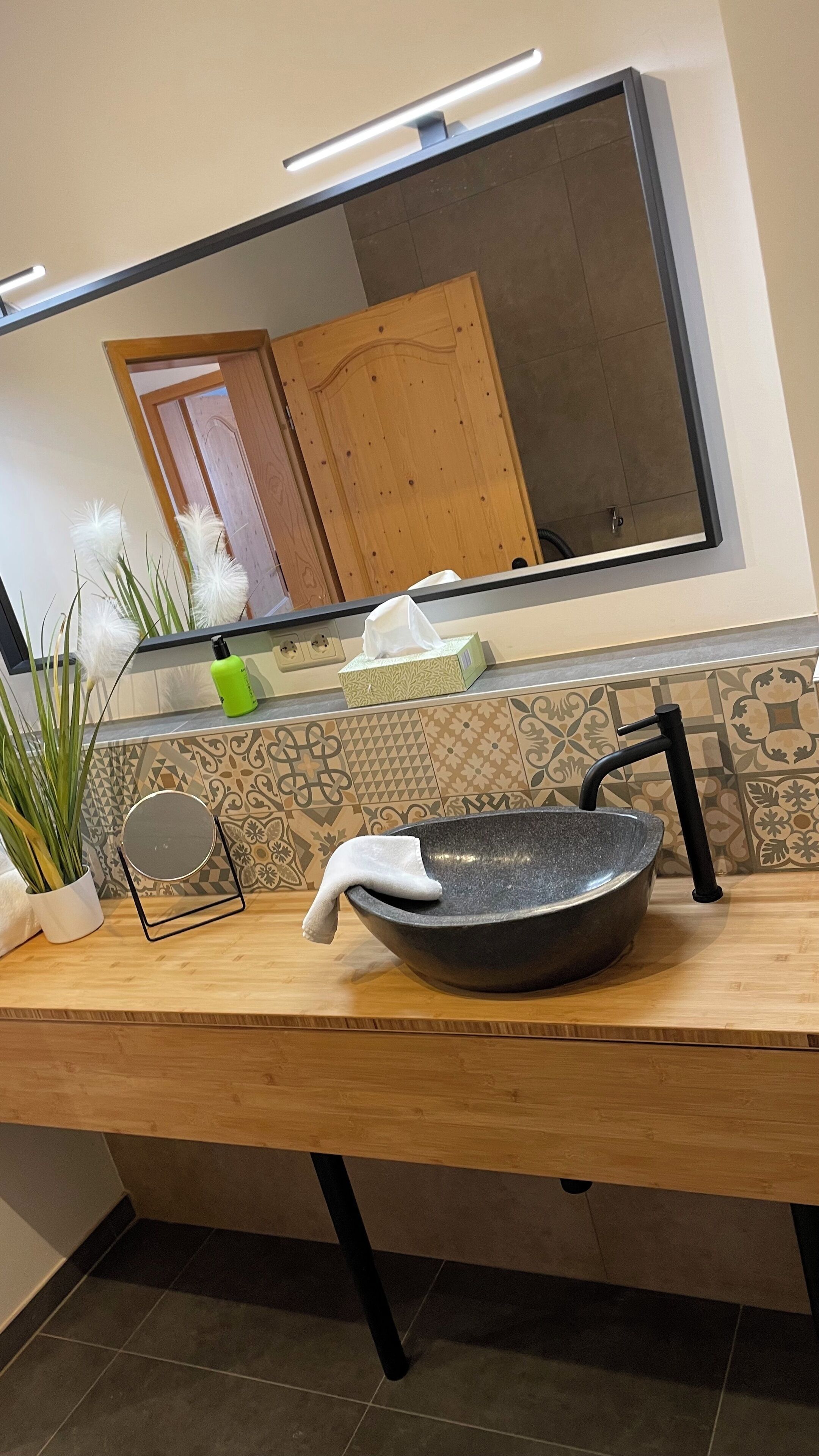 A bathroom with a wooden vanity, a large mirror, a black sink, and a potted plant.