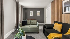 Un salon moderne avec un canapé gris, un canapé noir agrémenté d’un plaid jaune et un canapé vert avec un coussin noir. Deux tableaux encadrés ornent le mur, une plante est posée sur le sol et un luminaire est suspendu au plafond.