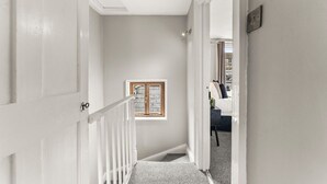 Un couloir avec une porte blanche, une petite fenêtre et un escalier menant à une pièce où se trouvent un lit et une chaise.