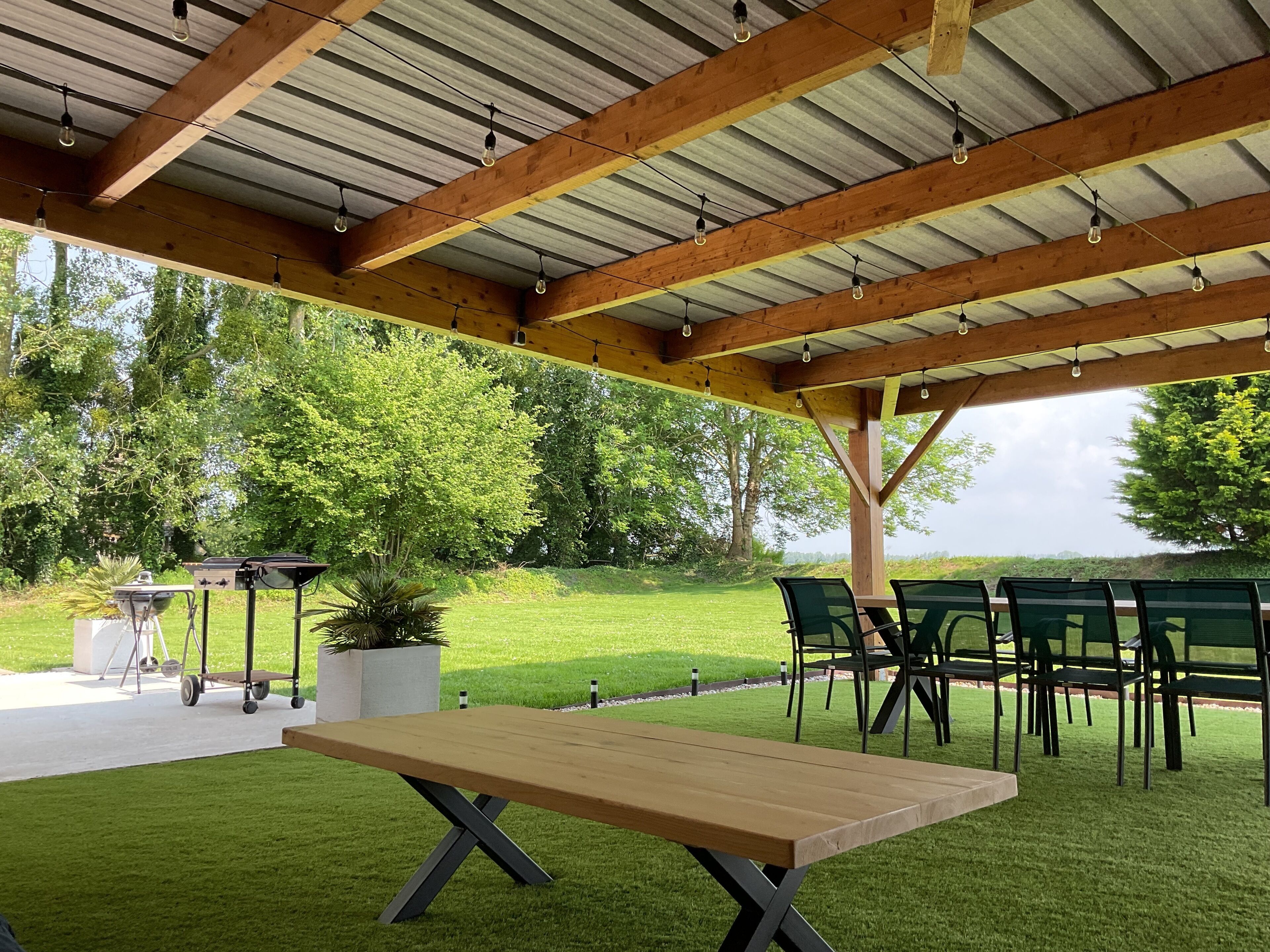 An outdoor patio with a wooden table and chairs, surrounded by greenery and trees.