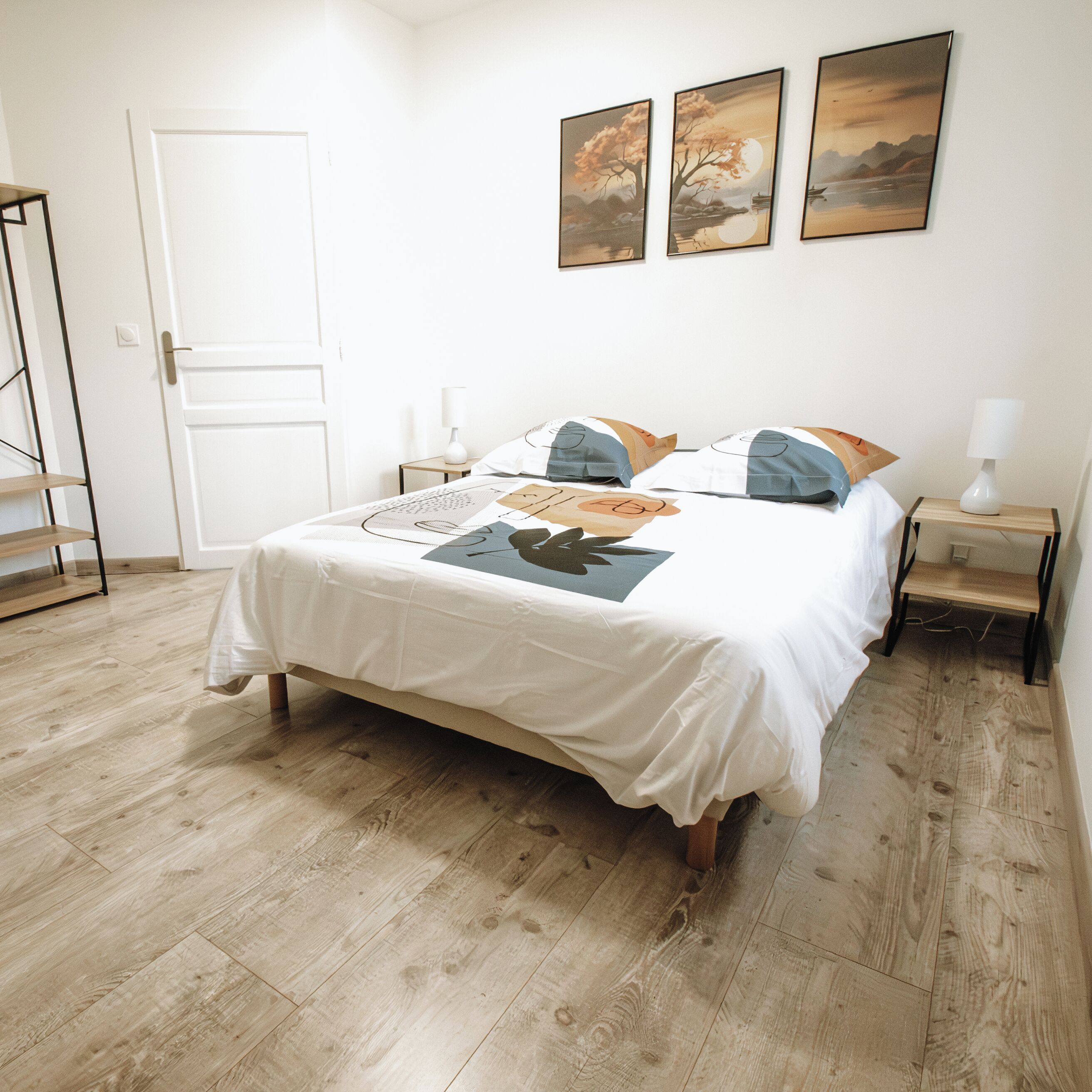 Un lit bien fait, recouvert d’une couvre-lit blanche et agrémenté d’un coussin au motif géométrique. Il y a trois tables de chevet, chacune surmontée d’une lampe blanche. La pièce est dotée d’un parquet et d’une porte blanche.