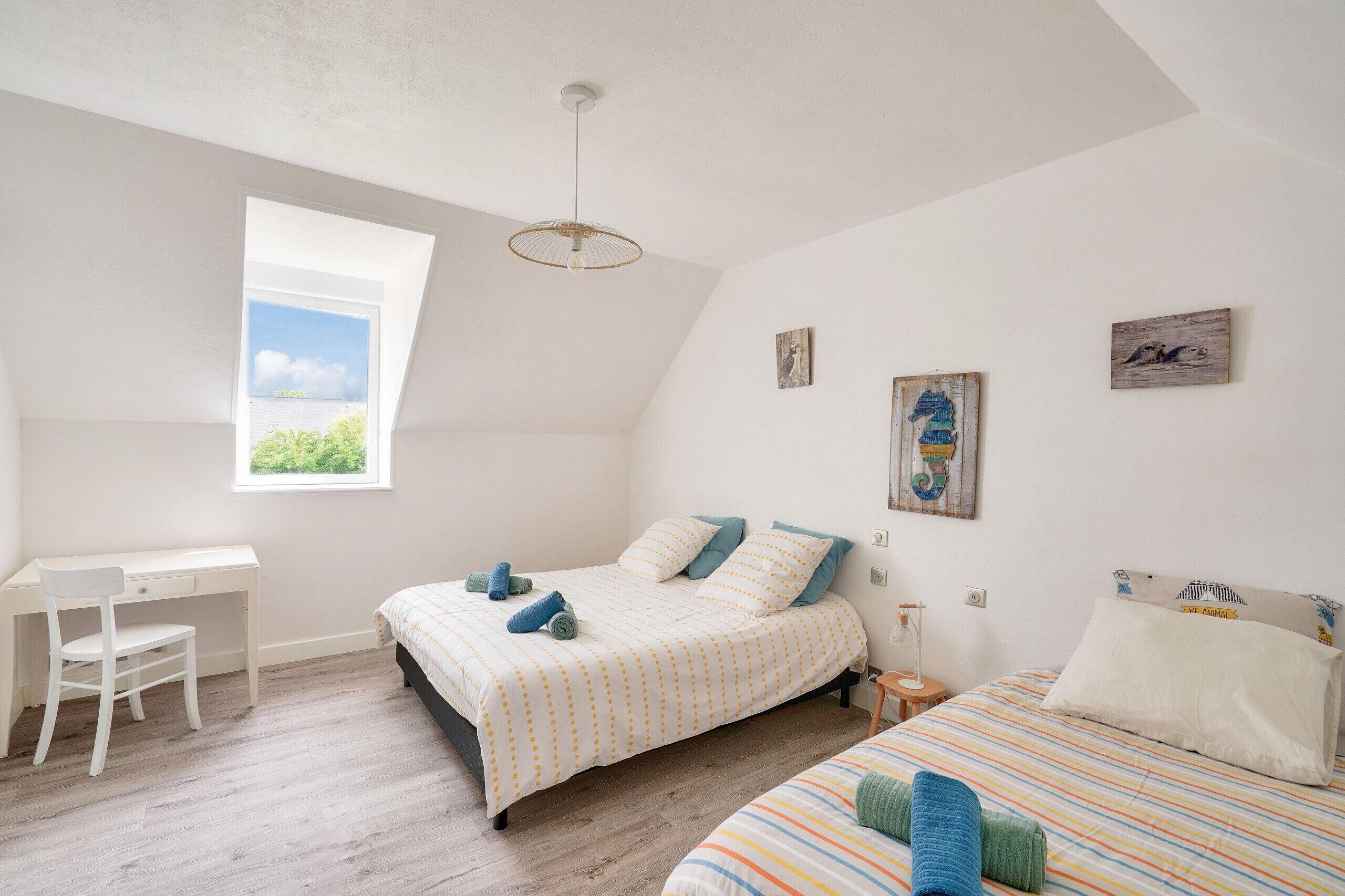 A bedroom with a desk, two beds, a window, and artwork on the walls.