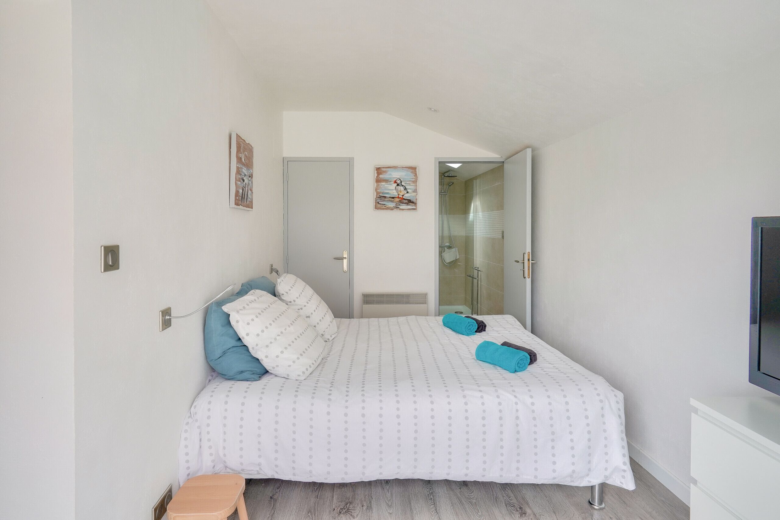 Quarto com cama, almofadas, um banquinho pequeno e uma televisão.