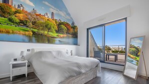 A bedroom with a large bed, a nightstand, a lamp, a window with a view of a park and cityscape, and a framed picture leaning against the wall.