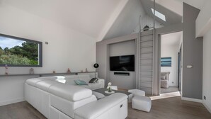 A modern living room with a sectional sofa, a flat-screen TV mounted on the wall, and a large window overlooking greenery.