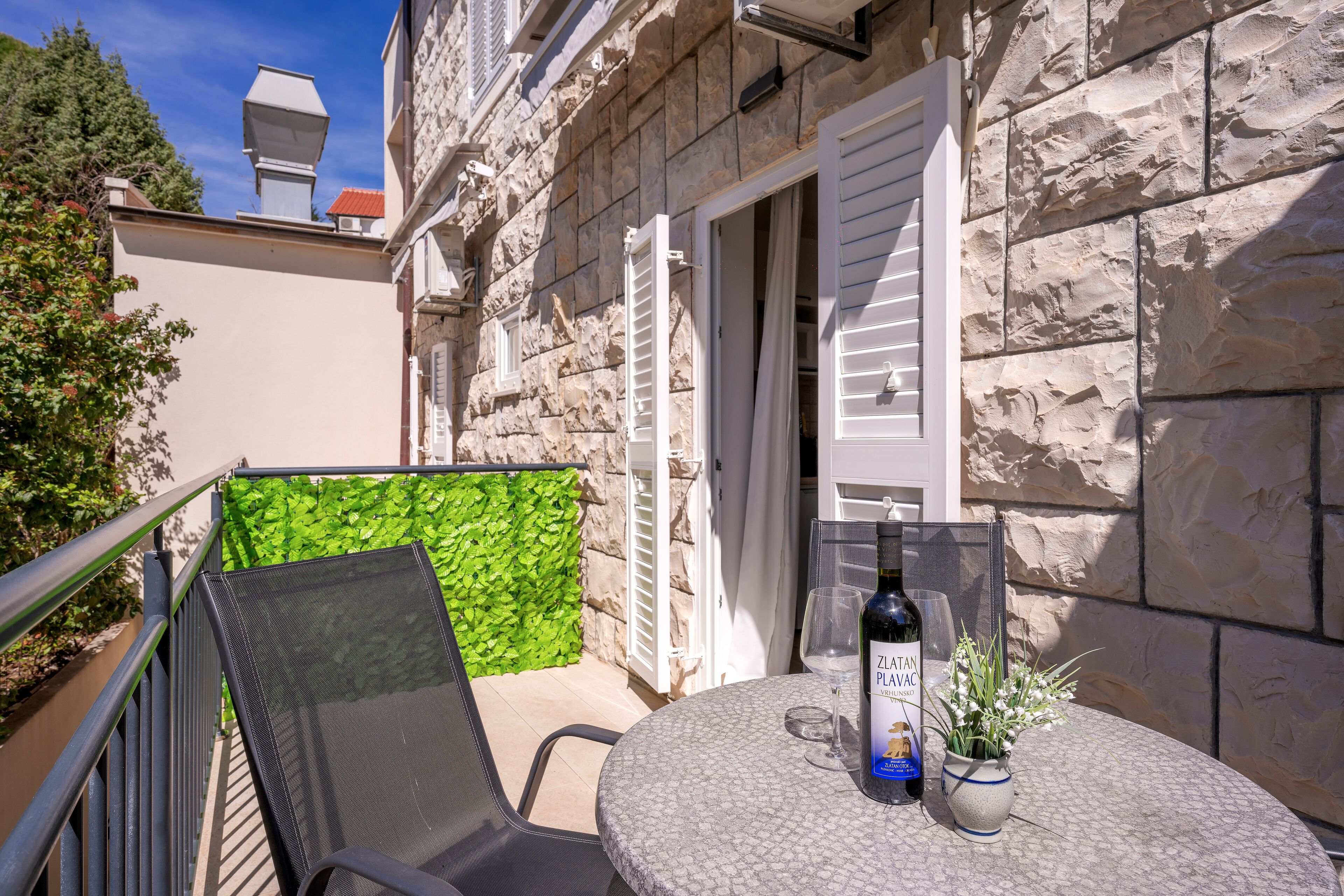 A stone building with a balcony, a table with a bottle of Zlatic Plavs wine, a glass, and a potted plant.