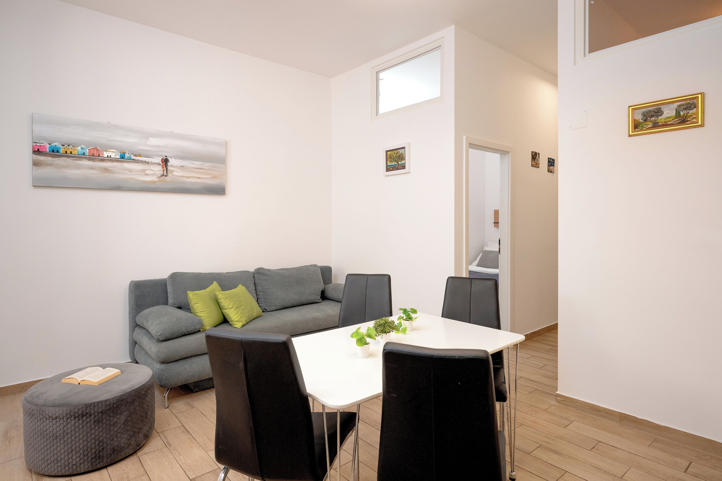 Un salon moderne avec un canapé gris, un petit pouf rond sur lequel repose un livre, une table blanche avec des chaises et un tableau accroché au mur.