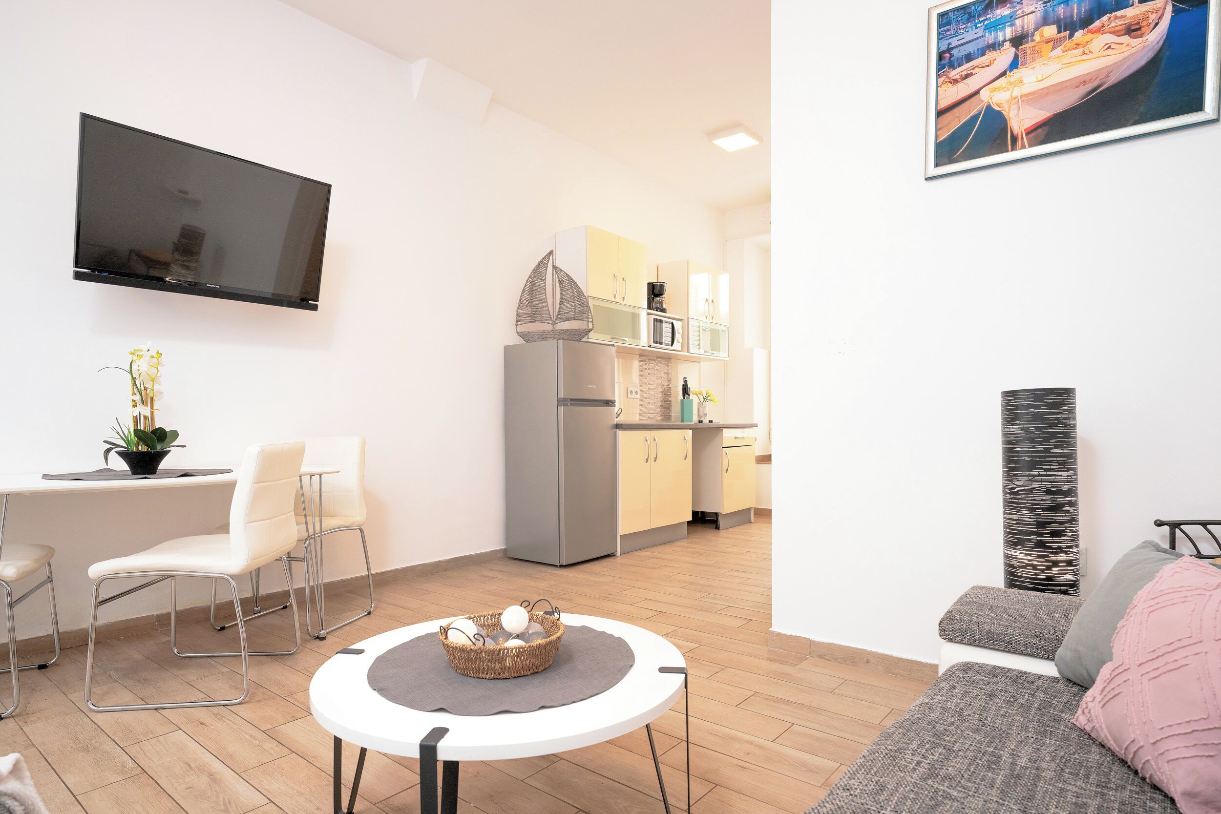 A modern living room with a flat-screen TV, a dining area with white chairs, a kitchen with a refrigerator and microwave, and a large window with a view of boats.