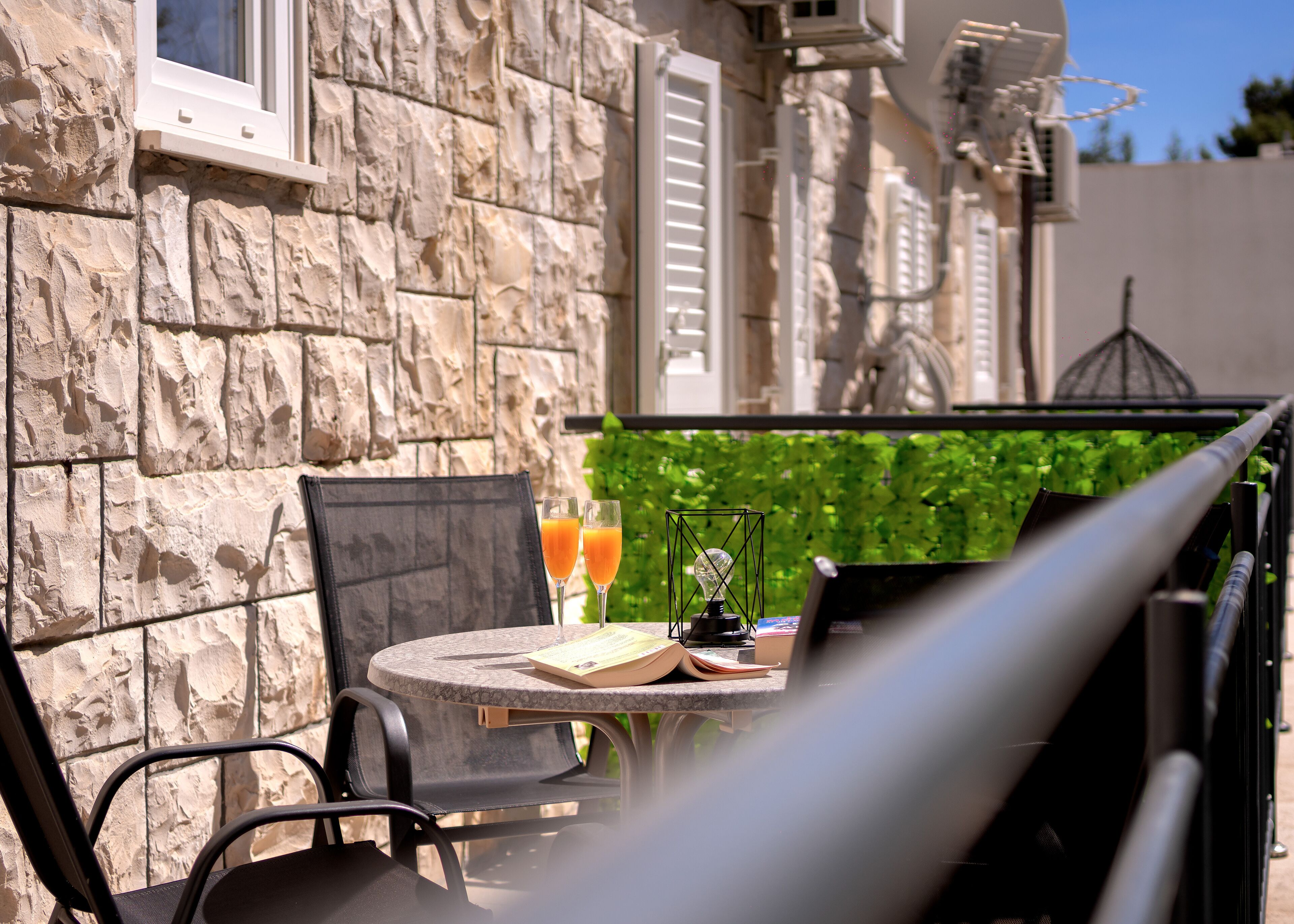 A stone building with a window and shutters, a table with two glasses of orange juice, a chair, and a decorative light fixture.