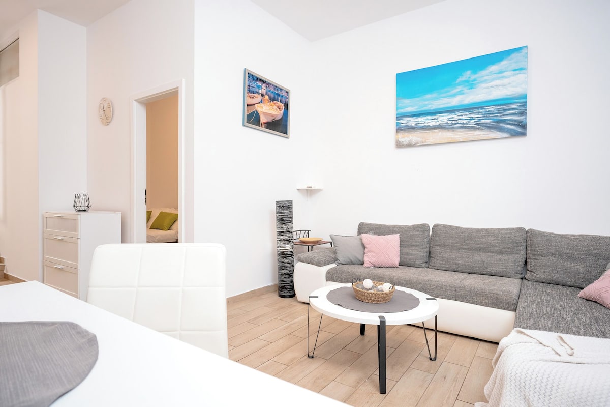 A modern living room with a grey sofa, a white armchair, and a round coffee table. There are two paintings on the wall, one depicting a cityscape and the other a beach scene. A white dresser and a lamp are also visible.