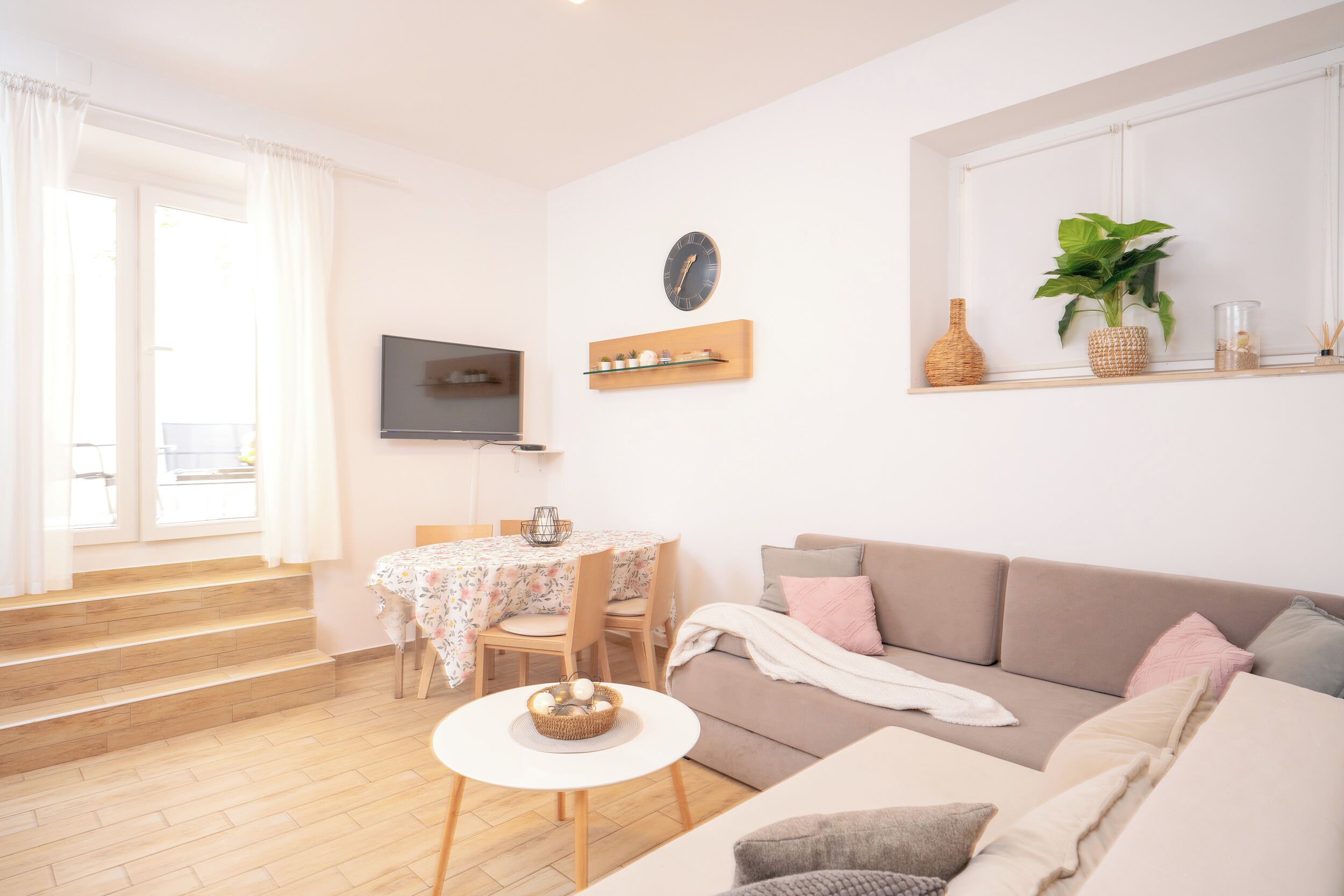 Un salon comprenant un canapé, une table à manger avec des chaises, un téléviseur fixé au mur et une fenêtre avec des rideaux.