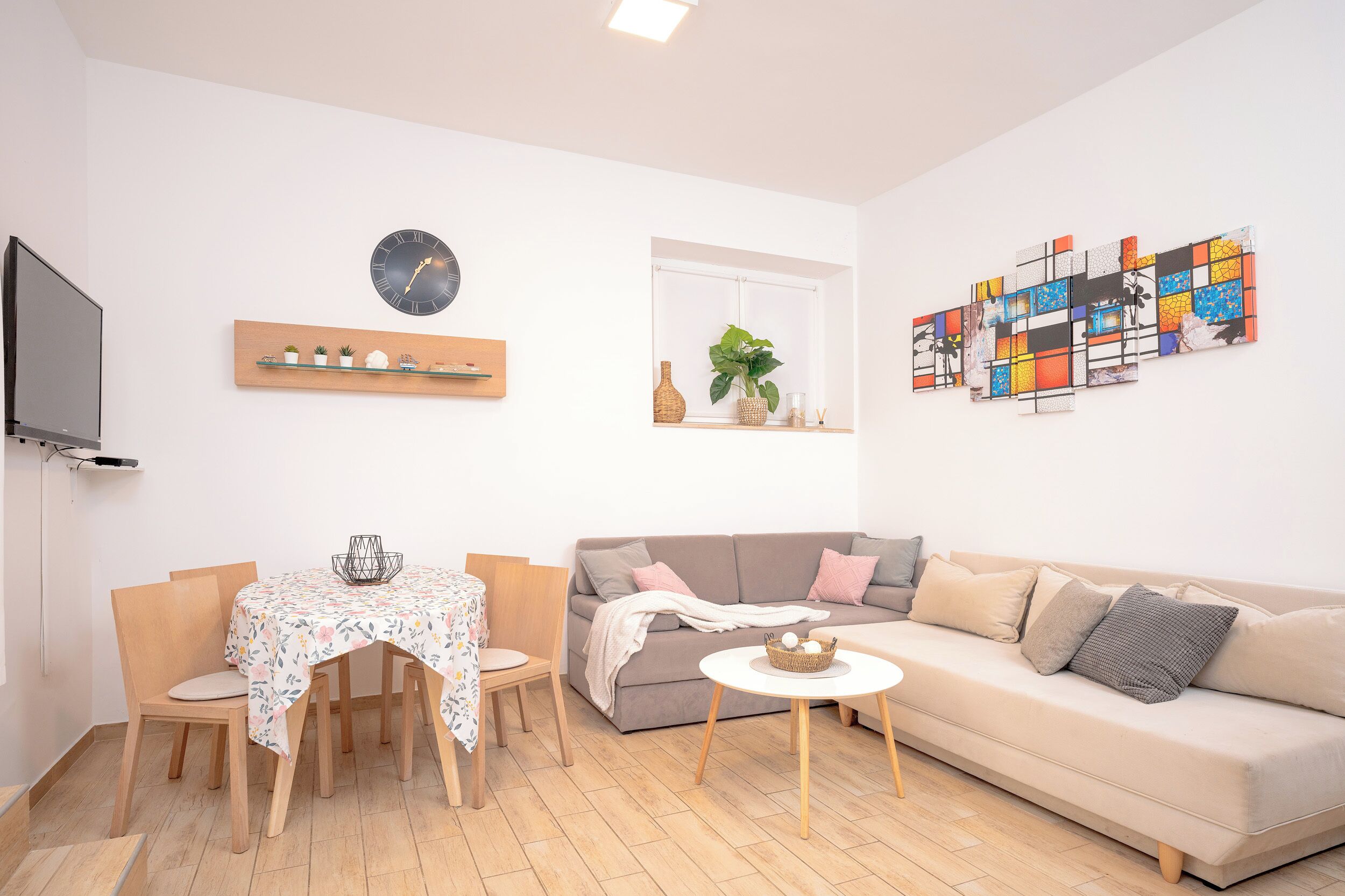Un salon moderne comprenant un canapé, une table à manger avec des chaises, un téléviseur, une horloge murale et un tableau abstrait aux couleurs vives.