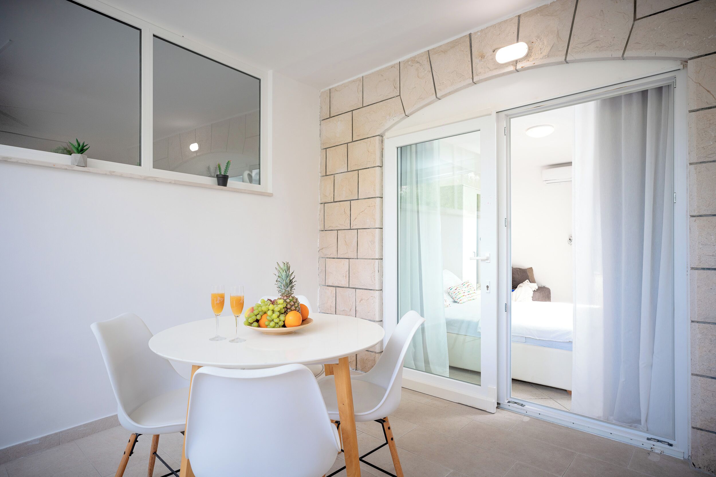 A dining area with a round table, white chairs, and a fruit bowl. There are potted plants on the windowsill and a sliding glass door leading to a bedroom.