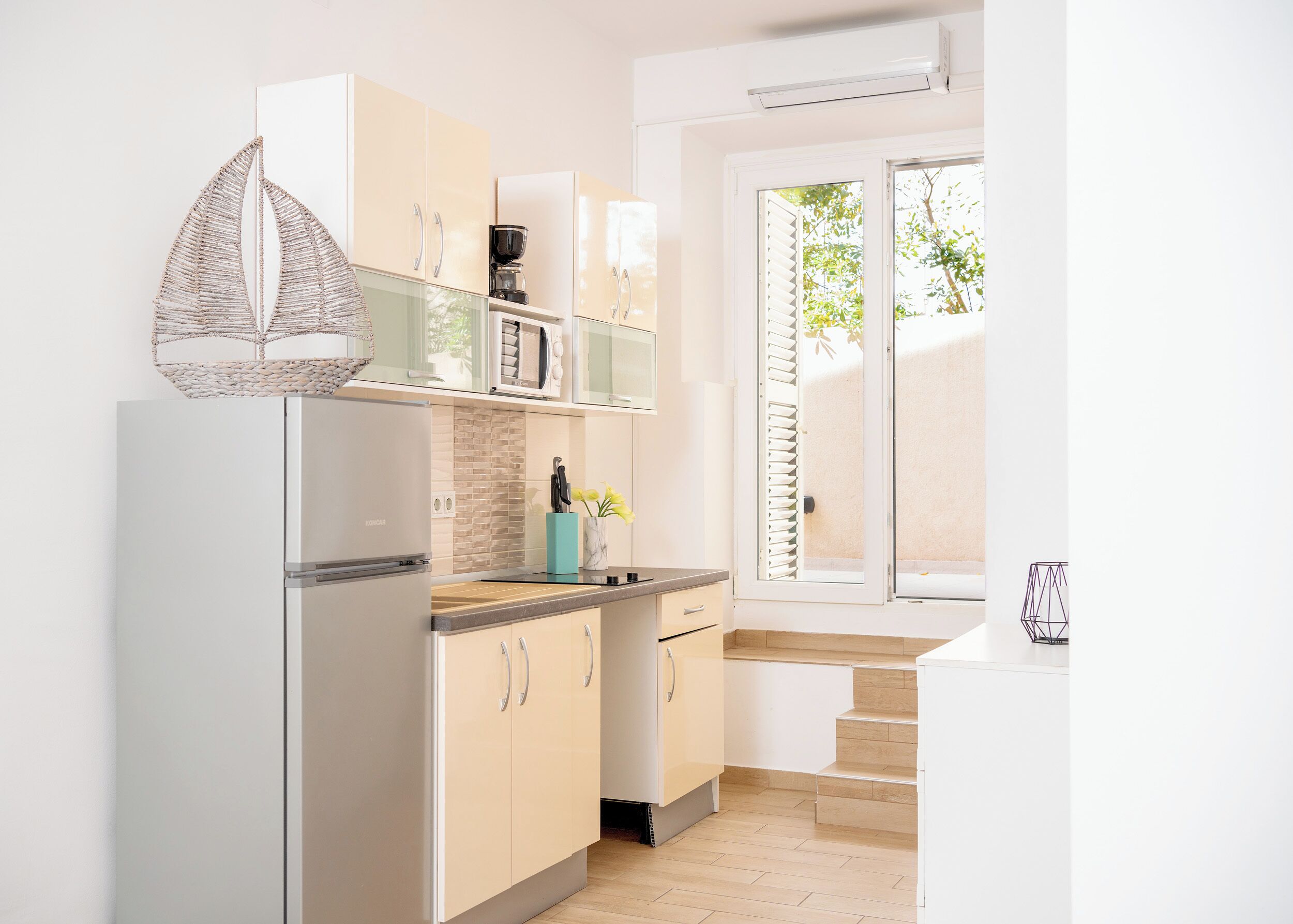 Una cocina moderna con armarios blancos, un microondas integrado y un refrigerador. Hay una ventana con persianas, un fregadero y un jarrón con flores en la encimera.