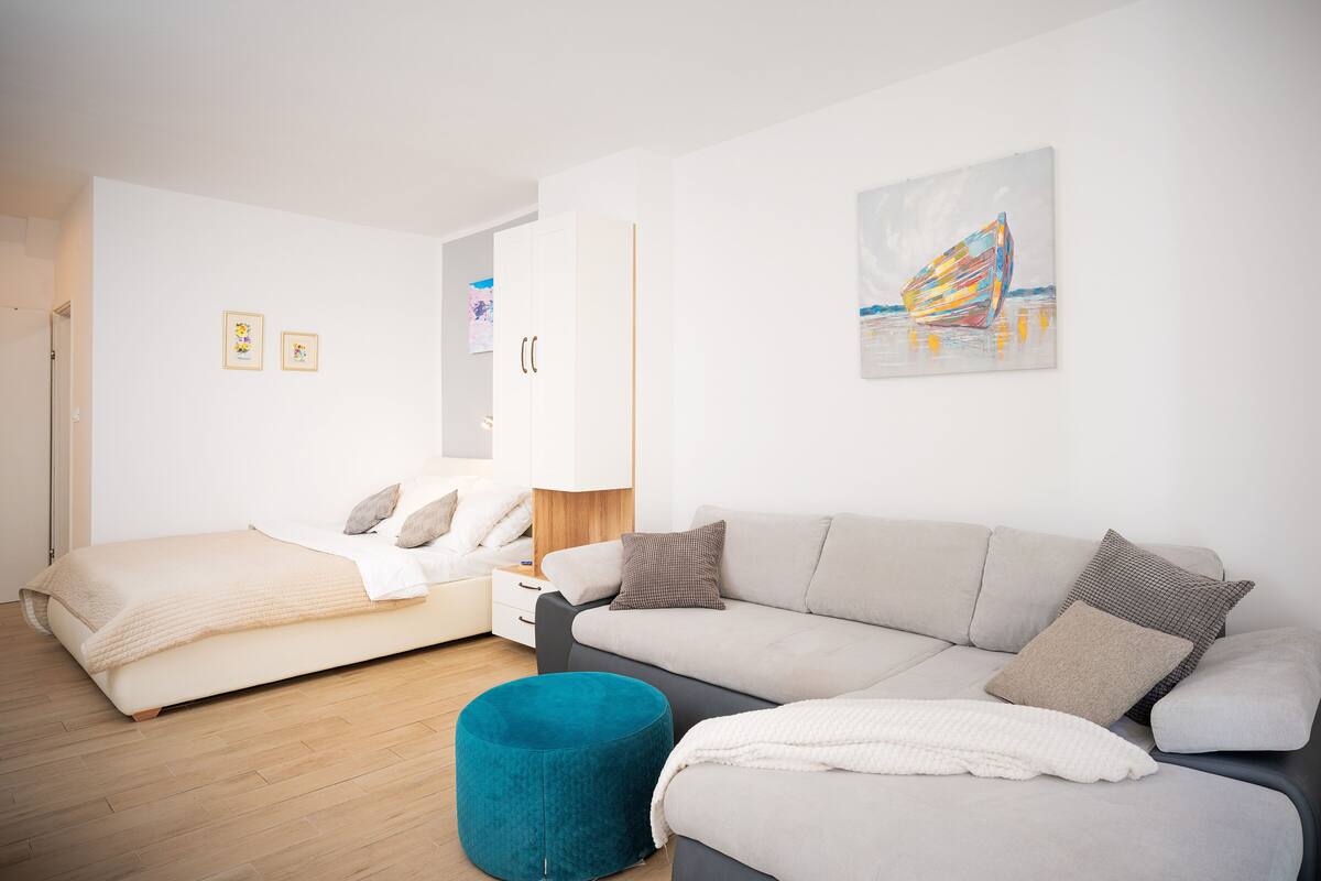 A modern, well-lit bedroom with a bed, a sofa, and a blue ottoman. There are two framed pictures on the wall and a colorful abstract painting above the sofa.