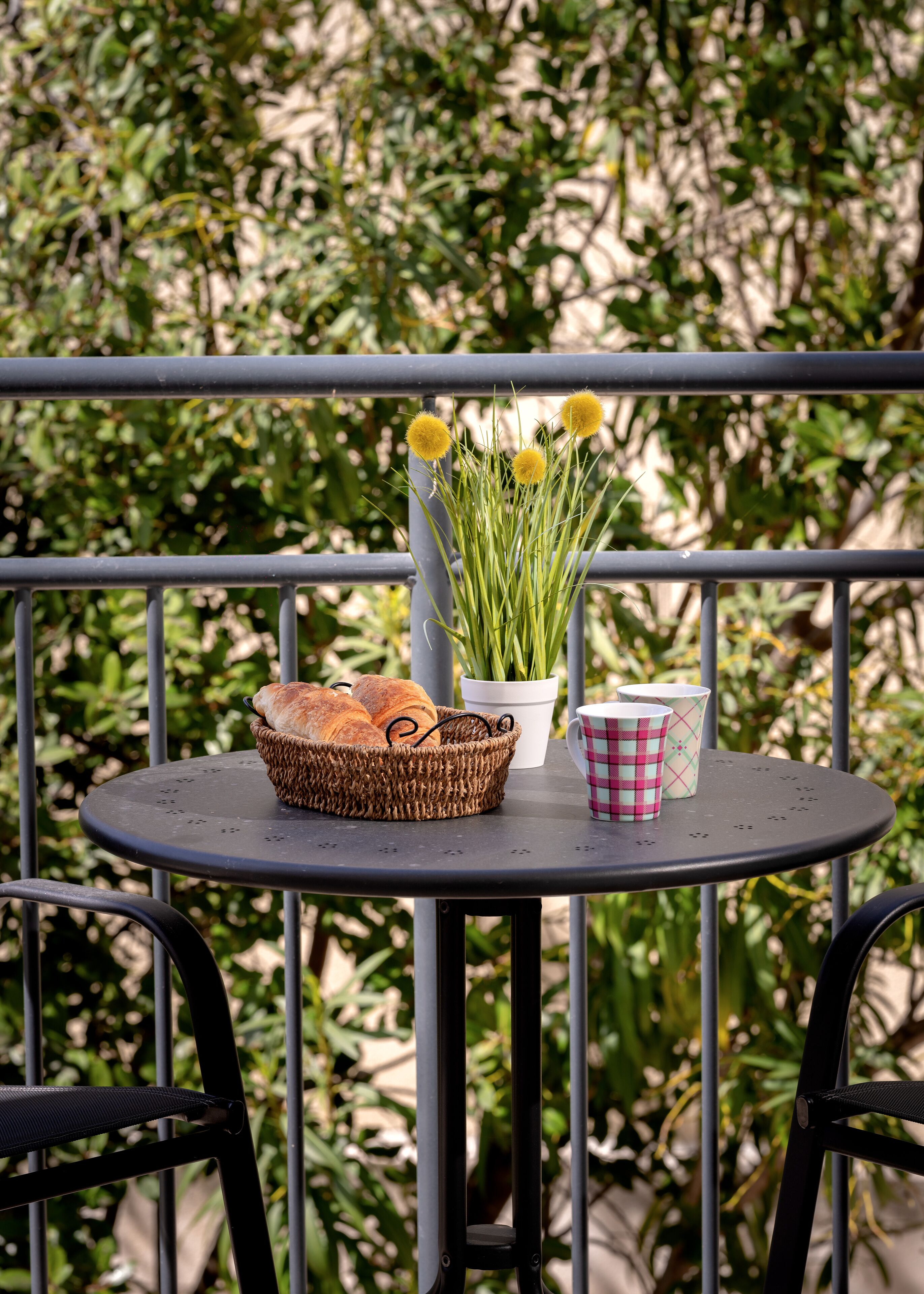 Une table ronde en métal sur laquelle se trouve un panier en osier contenant du pain, un pot blanc avec des plantes vertes et une tasse à carreaux.