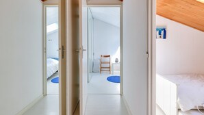 A bedroom with a white bed, a wooden ceiling, and a view of another room with a chair and a table.