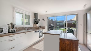 A modern kitchen with white cabinets, a wooden countertop, and a large island. There is a window above the sink and a sliding glass door leading to a backyard. - Family house 400m from the sea for 8, Pornic (Pornic)