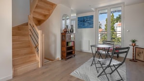 A modern interior with a wooden staircase, a dining area with a table and chairs, a bookshelf, and a large window offering a view of the outside. - Family house 400m from the sea for 8, Pornic (Pornic)