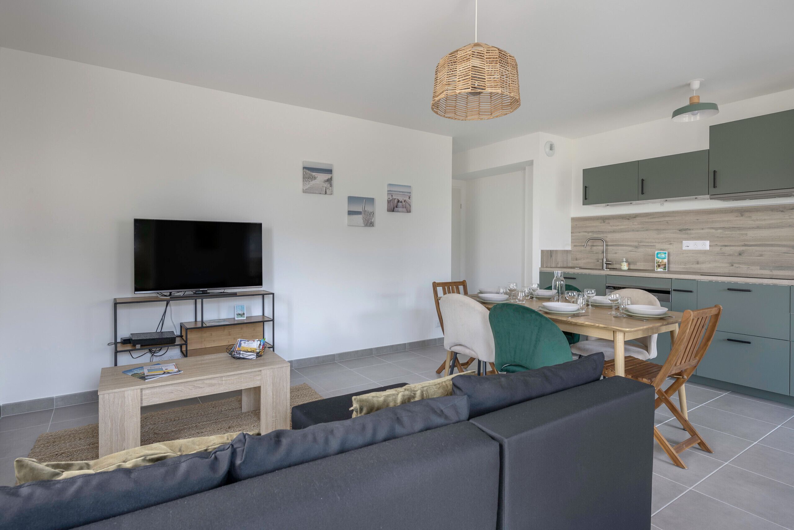 Una sala de estar moderna con un televisor de pantalla plana, una mesa de centro de madera y un sofá gris. Hay un área de comedor con una mesa preparada para dos personas, y una cocina con armarios verde claro.