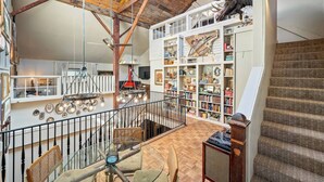 A spacious loft with exposed wooden beams, a mezzanine level with a bookshelf, and a staircase with patterned carpet.