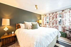 A hotel room with a large bed, two bedside tables with lamps, floral curtains, and a green suitcase on the floor.