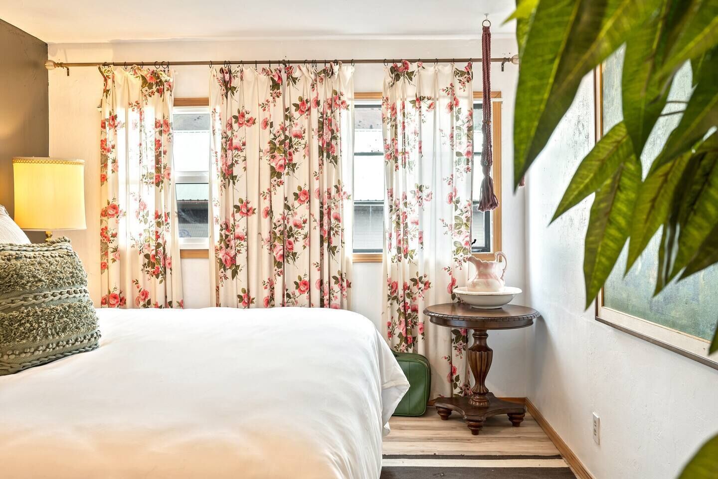 Une chambre d’hôtel avec un lit, une lampe, une table avec une carafe et des rideaux à motifs floraux.
