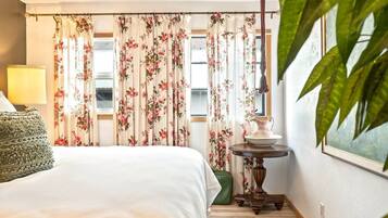 Une chambre d’hôtel avec un lit, une lampe, une table avec une carafe et des rideaux à motifs floraux.